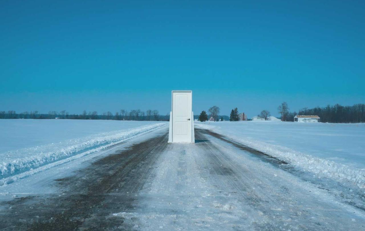 A theatrical set door stands in the middle of a snowploughed road between fields of snow under a blue sky.