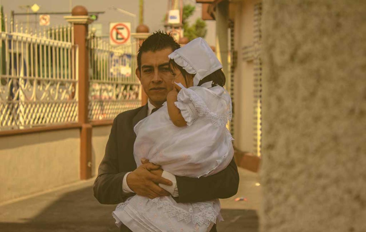 A father wearing a suit carries his child who is dressed for a christening in white.