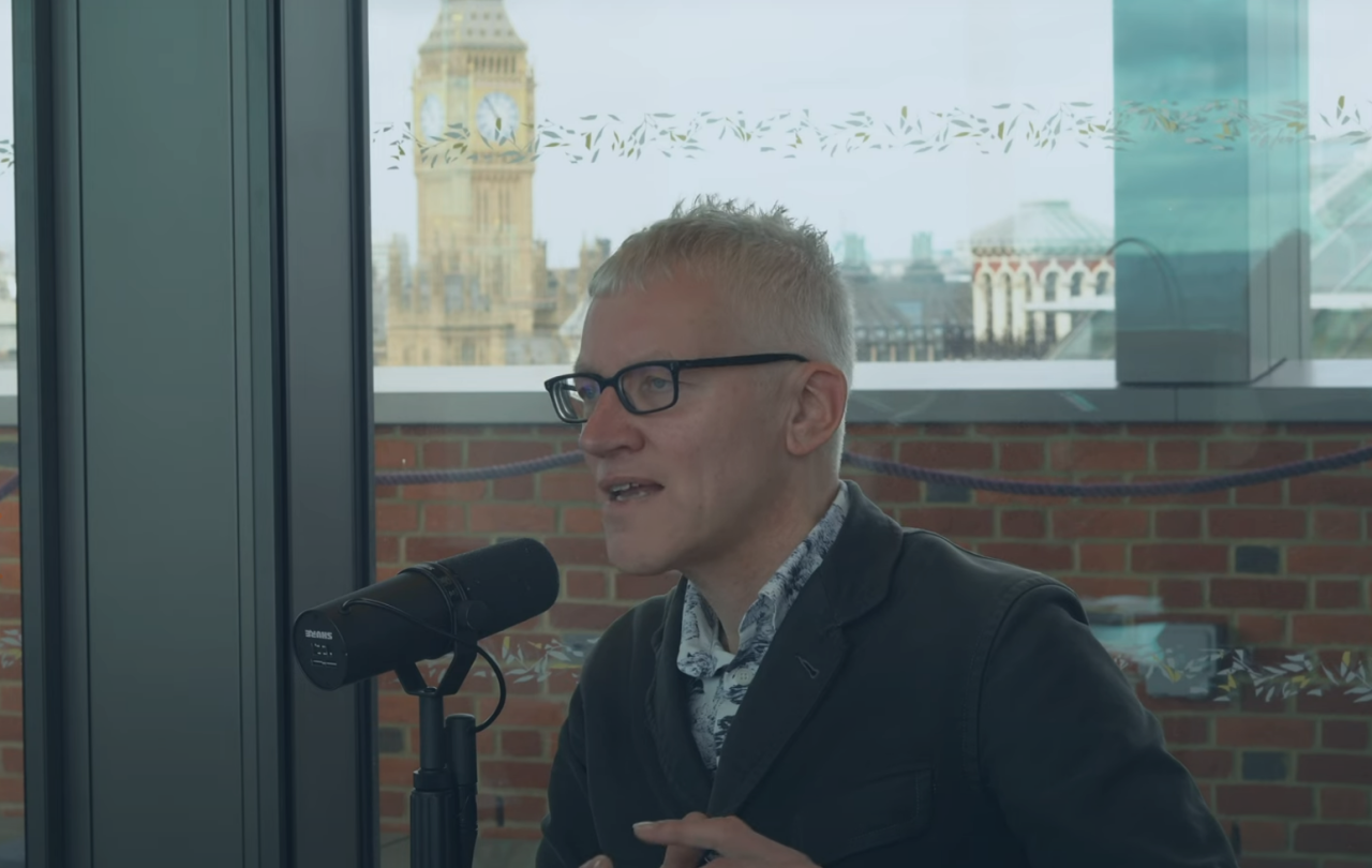 A man sits at a table speaking into a microphone but looks into the distance. Behind him, through the window and beyond a wall is Big Ben