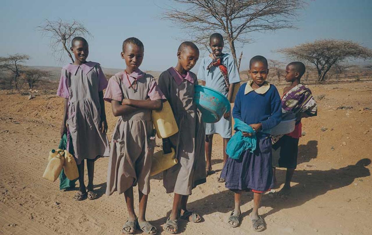 Girls stand in the northern Kenyan scrubland holding water bottles.