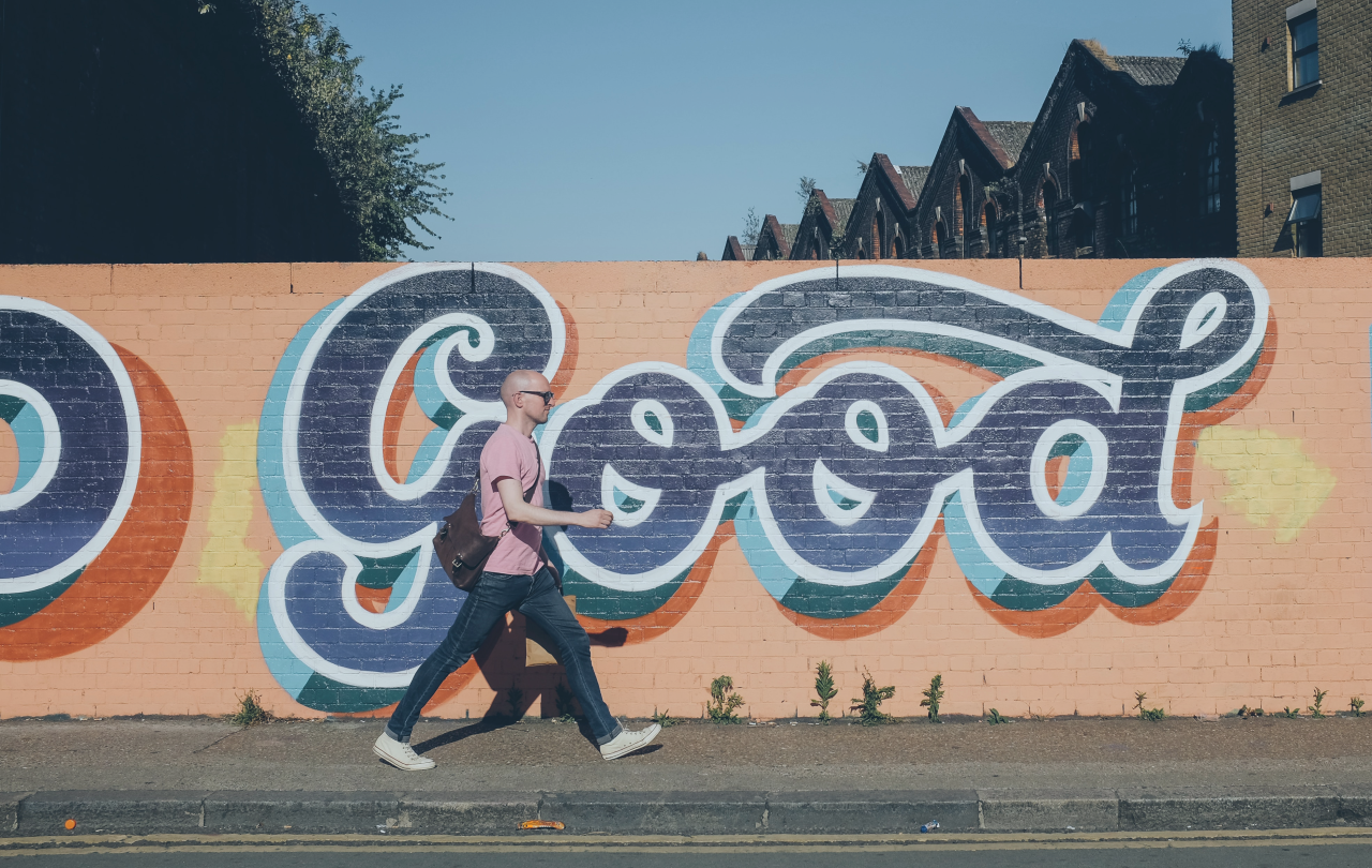 A man walks along a street past a orange wall with a huge 'Good' written in cursive script on it.