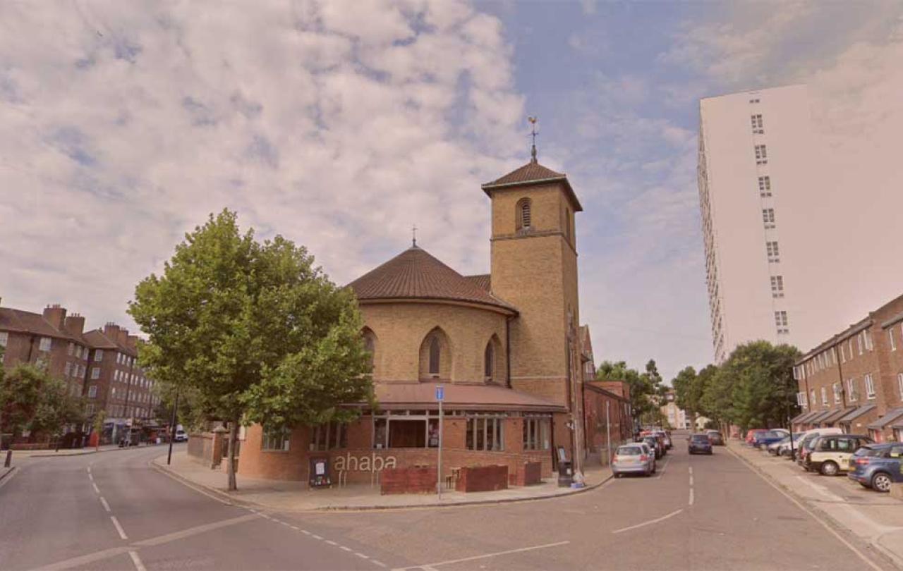 A neo-Romanesque church sits at the acute corner of two roads. To its side a tower block rises over a row of low-rise flats.