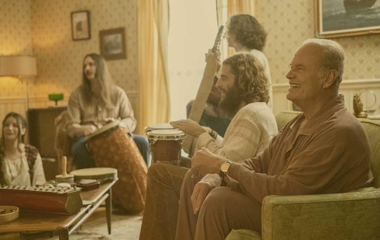 A group sit in a lounge playing musical instruments while the man closest to the camera laughs.