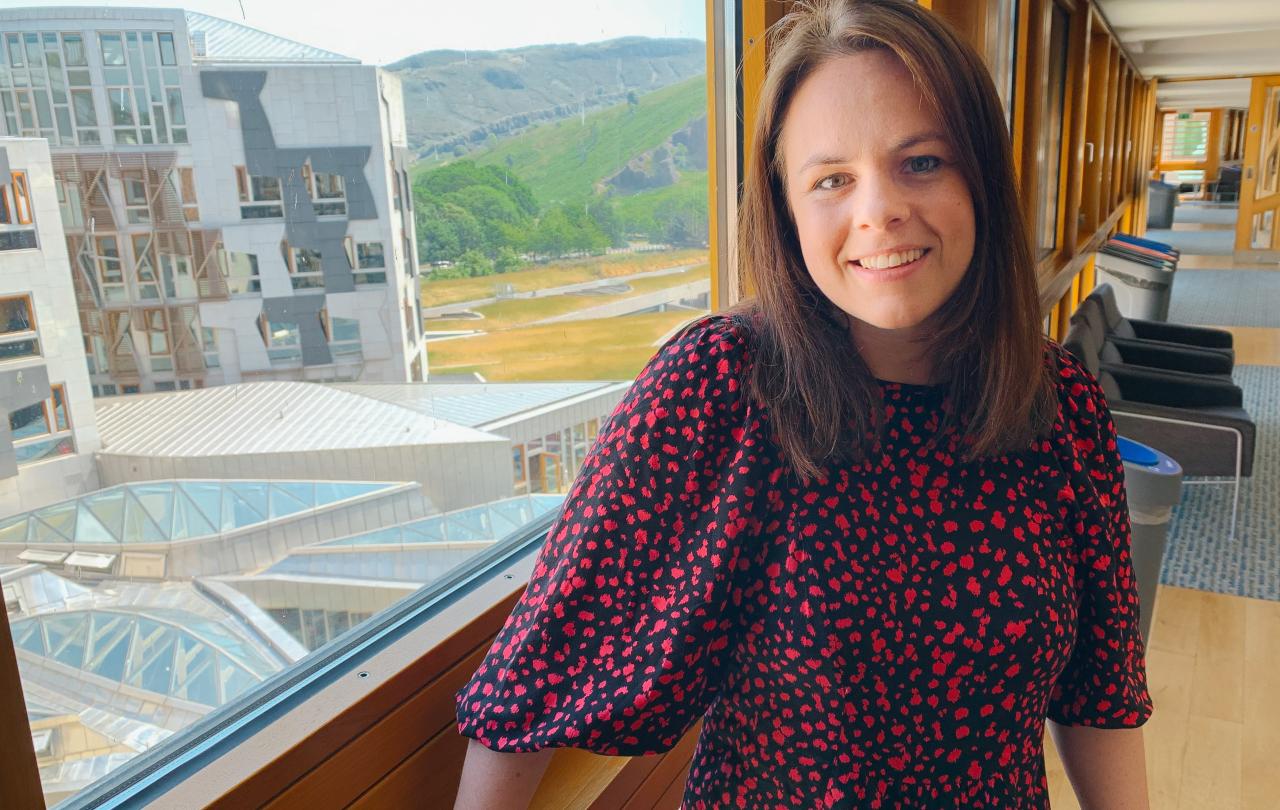 A women stands beside in a corridor beside a large window through which a wing of a building and a distant hillside can be see,