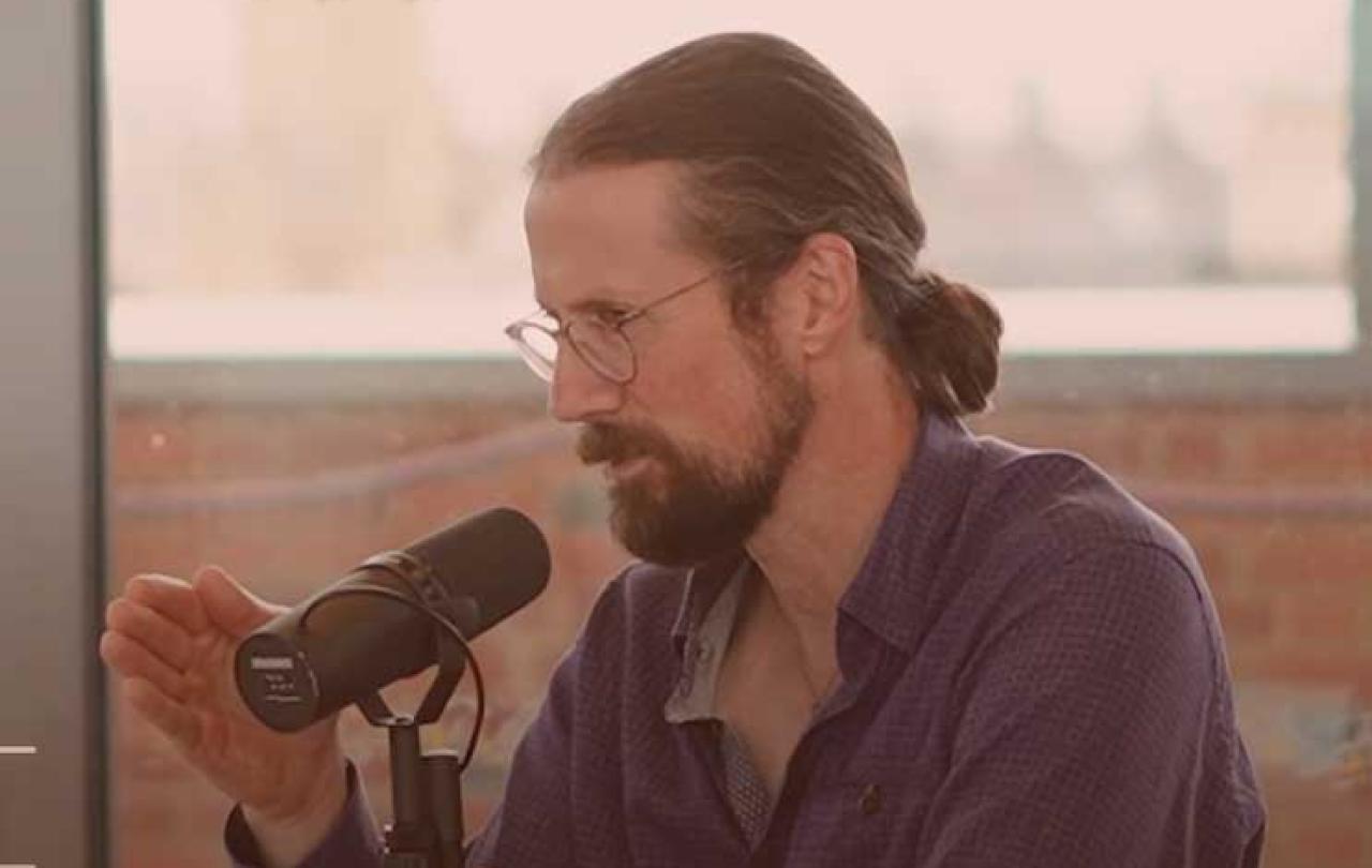 A sitting man speaks into a microphone will gesturing with one hand