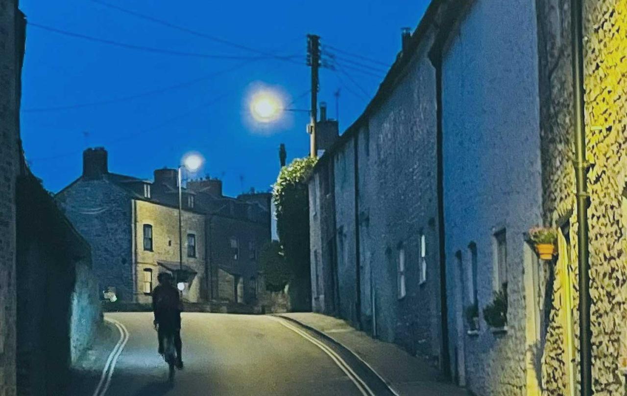 A cyclist ascends a village street between stone-built houses as twilight turns to night