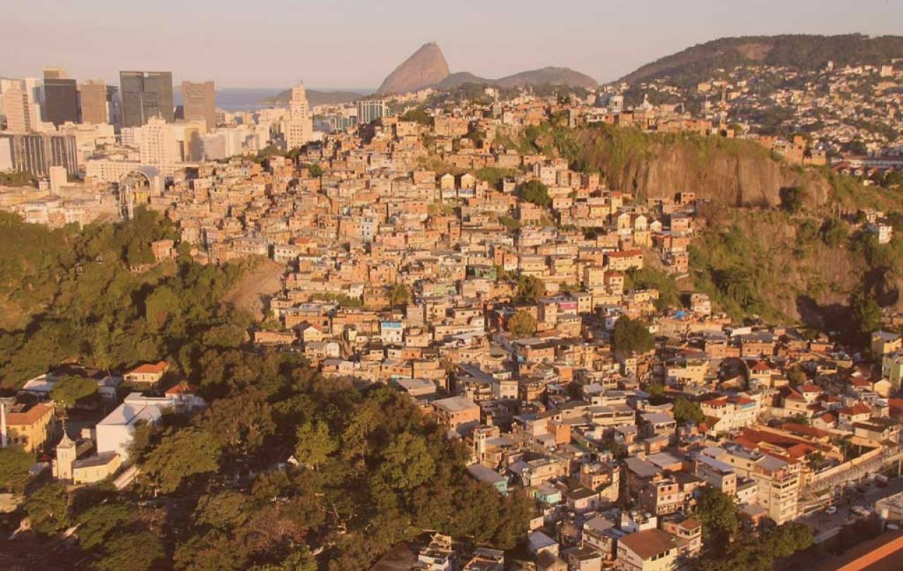 an aerial view of a shantytown on a steep hill side in Rio, Sugerloaf Mountain is visible in the distance 