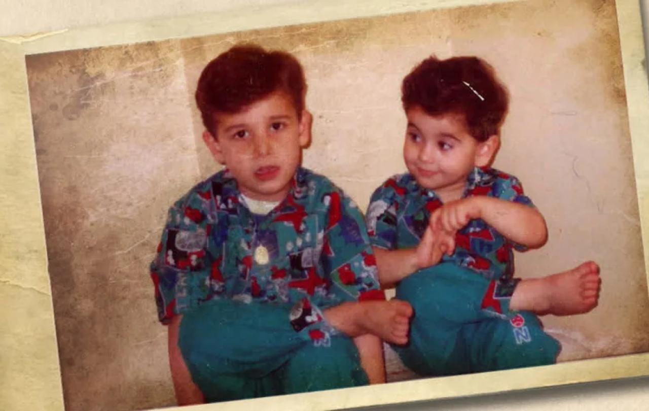 Two young brothers sit next to other, the younger looks to the elder.