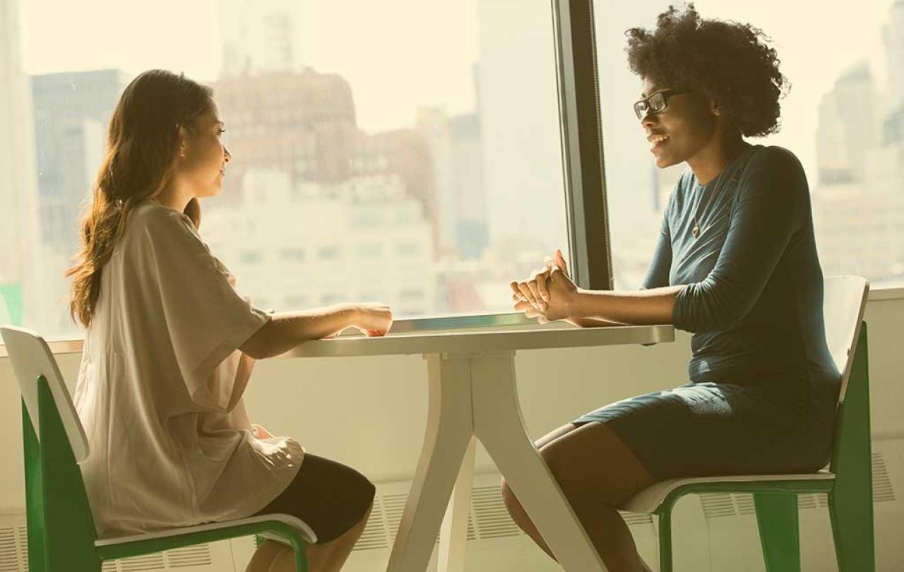 Two people sit at a table with their hands resting on top of it. One speaks to the other