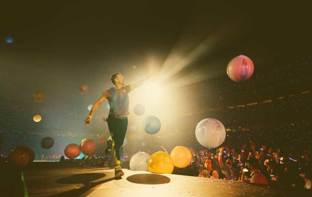 A singer struts a stage pointing to the spotlight as coloured orbs float down.