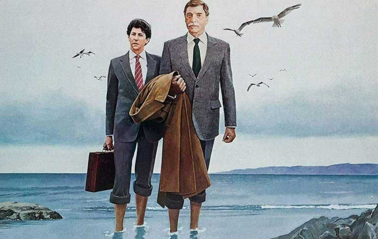 Two business men in suits hold coats and briefcases, stand in the sea with their trousers rolled-up above their ankles