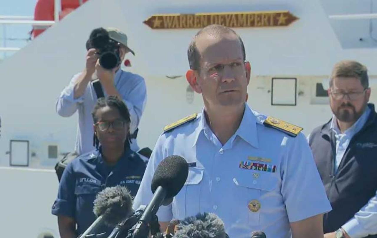 A Coast Guard officer gives a press conference while looking grim-faced. Others look on.