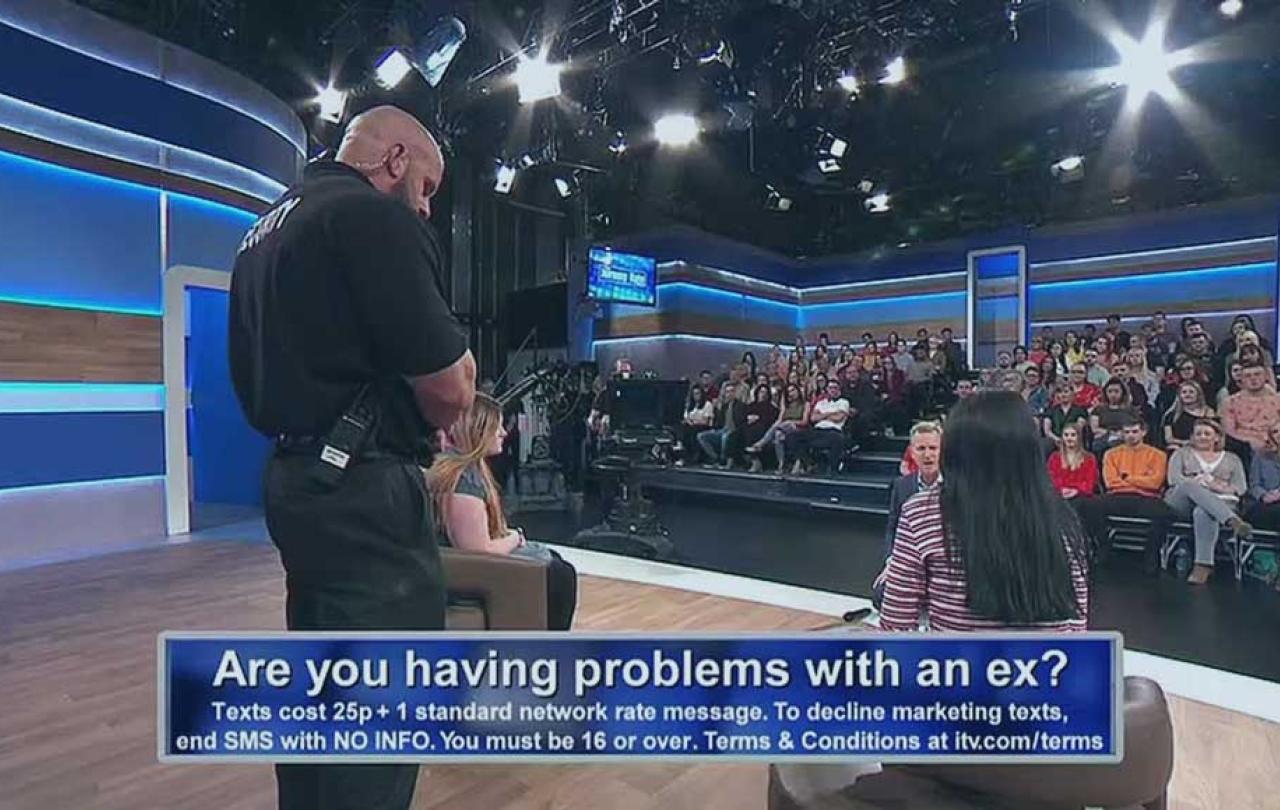 A view across a talk show TV set showing a security guard standing between two guest while the host talks to them. An audience looks on.