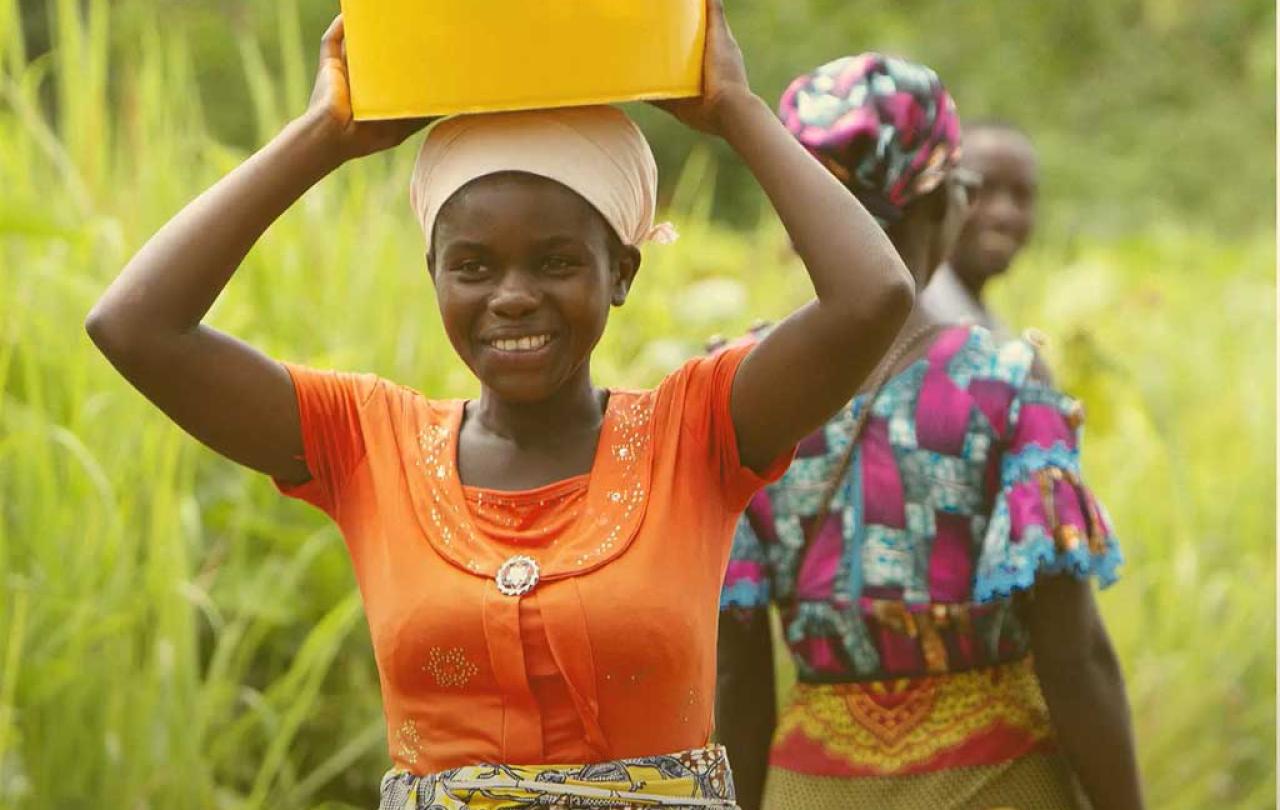 A woman carries a plastic bucket on her head and held by raised arms and hands.