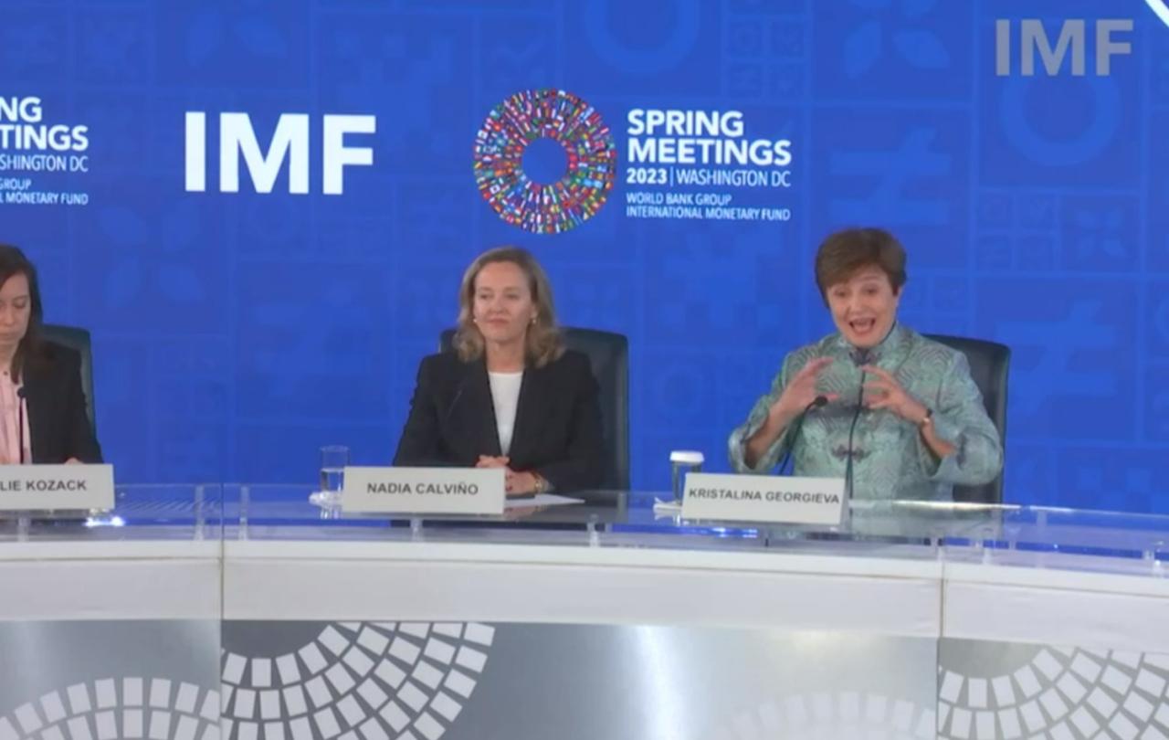 Two women sit behind a press conference desk against a backdrop, one listens as the other speaks and  gestures.
