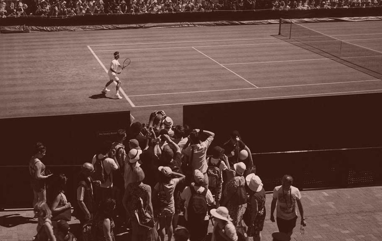 A tennis player stands ready to return a shot, while a phalanx of photographers crowd round a court-side opening to take a picture of him.