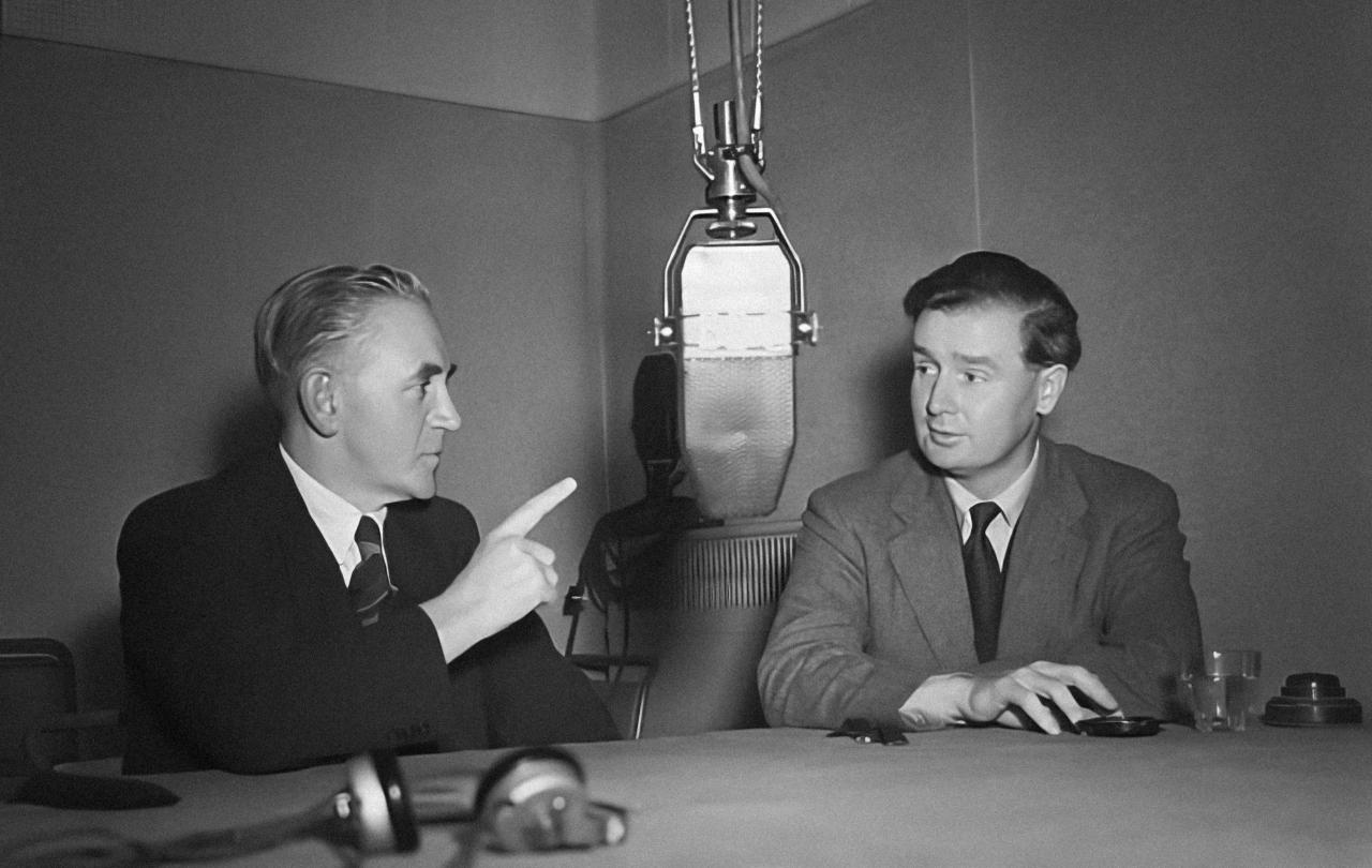 Two 1950's men un suits sit at a table dominated by a large hanging microphone. One points a raise hand and finger into the air. The other listens.