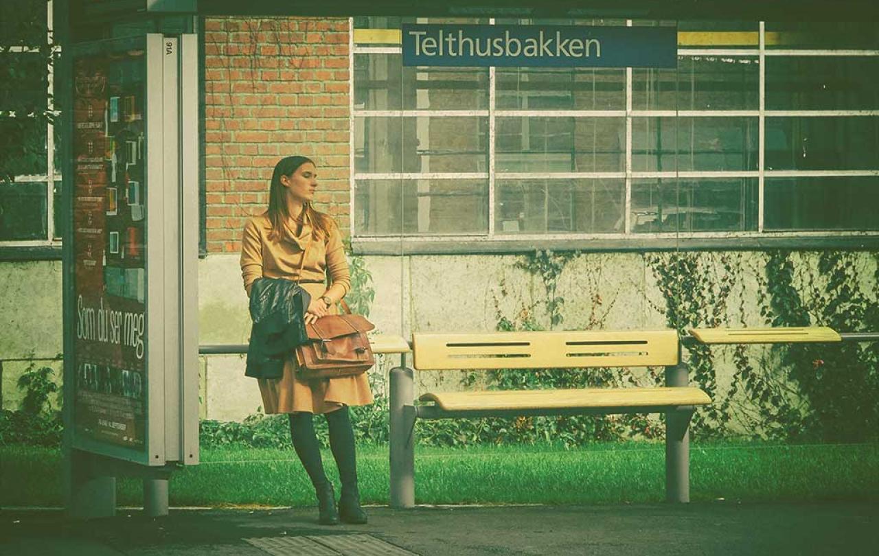 A woman leans against the glass of a bus shelter while waiting, she clasps a bag.