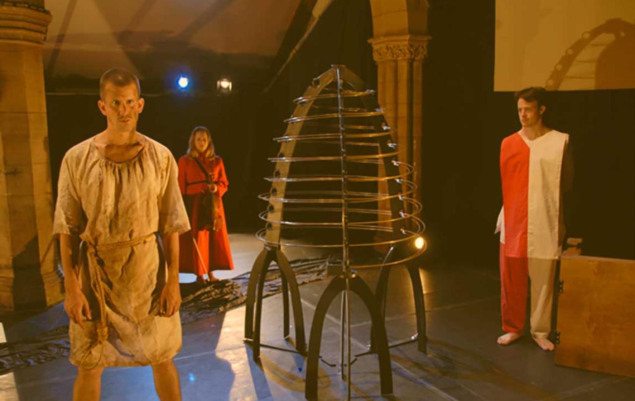 Three actors stand on a stage, in costume, surrounding a metal conical structure.