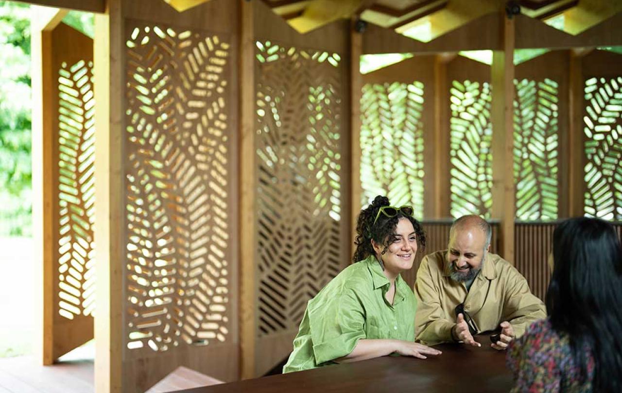 Around a table, against a backdrop of fret-cut wood, three people talk and listen to each other with great interest.