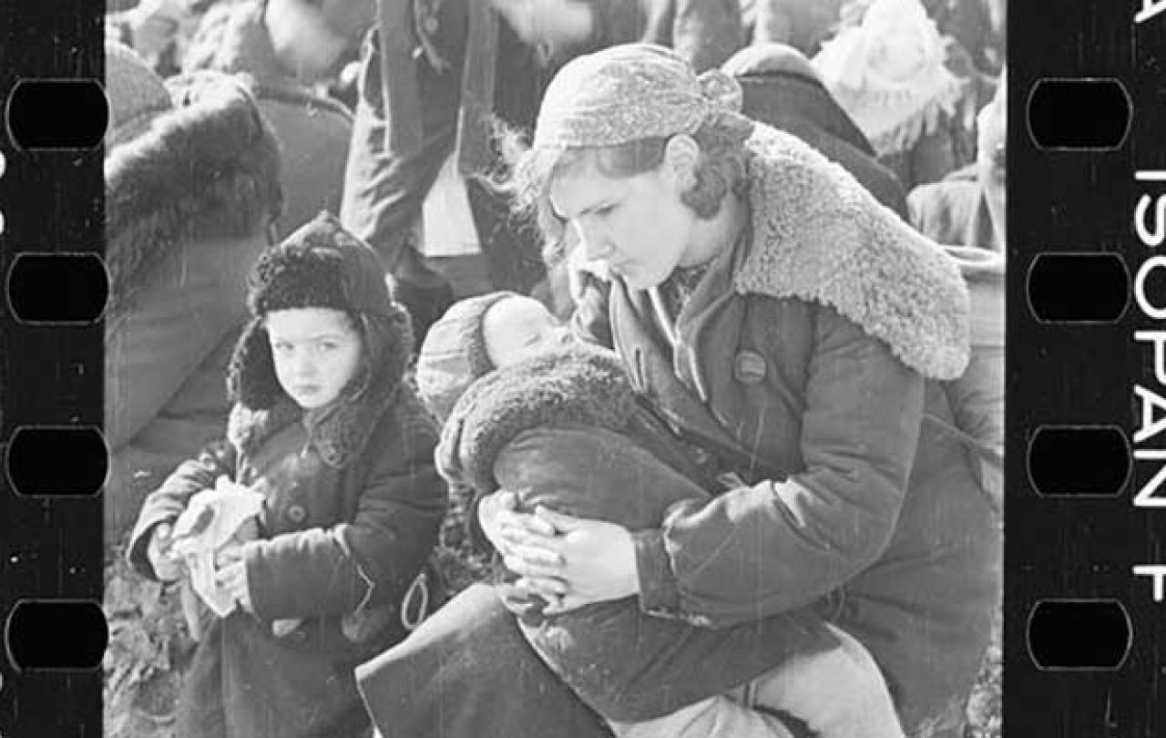 A mother cradles a child while another stands close by. They wear winter clothes of the 1940s and are amidst others waiting.