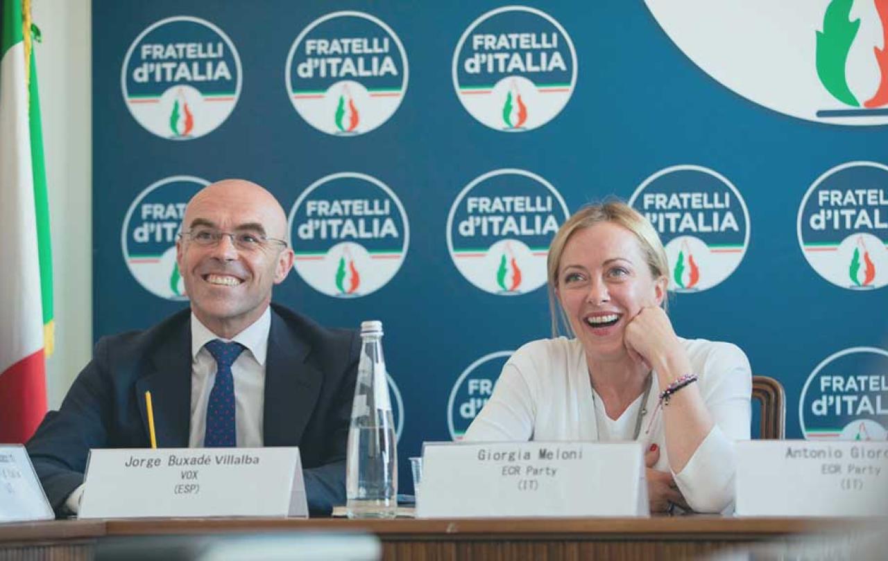 two politician site at a press conference desk and laugh, behind them is a backdrop of the political party's logo.