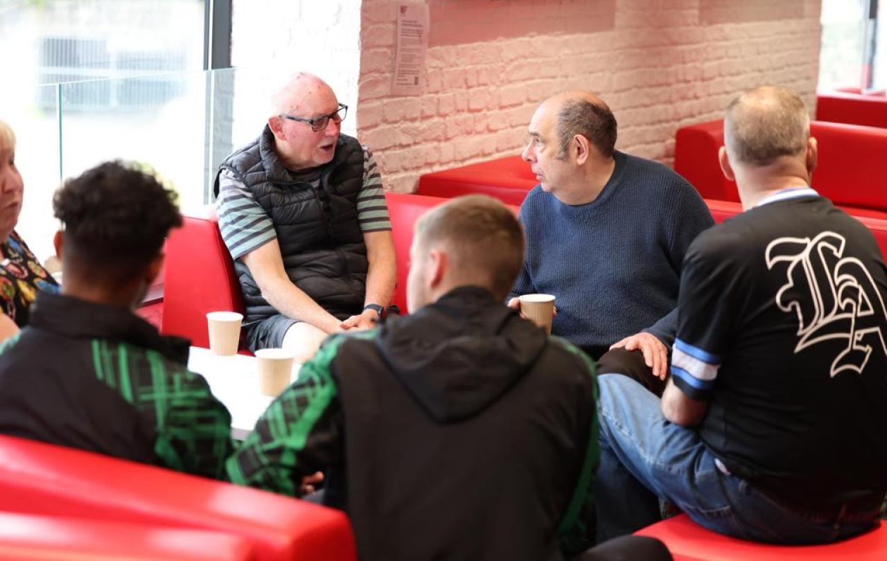 A group of old and young men sit on red sofas, listening to one of themselves.