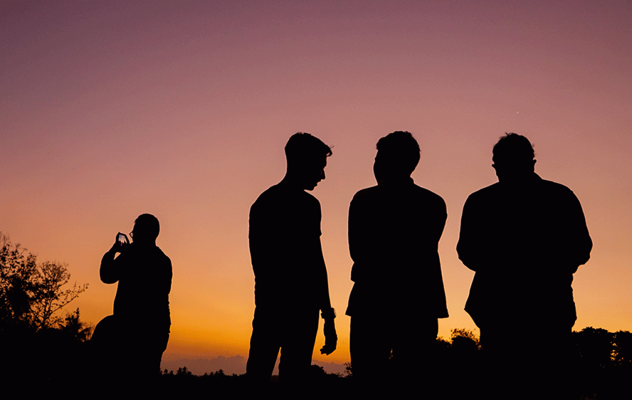 Four men stand silhoutted against a sunset, One stands apart on their phone.