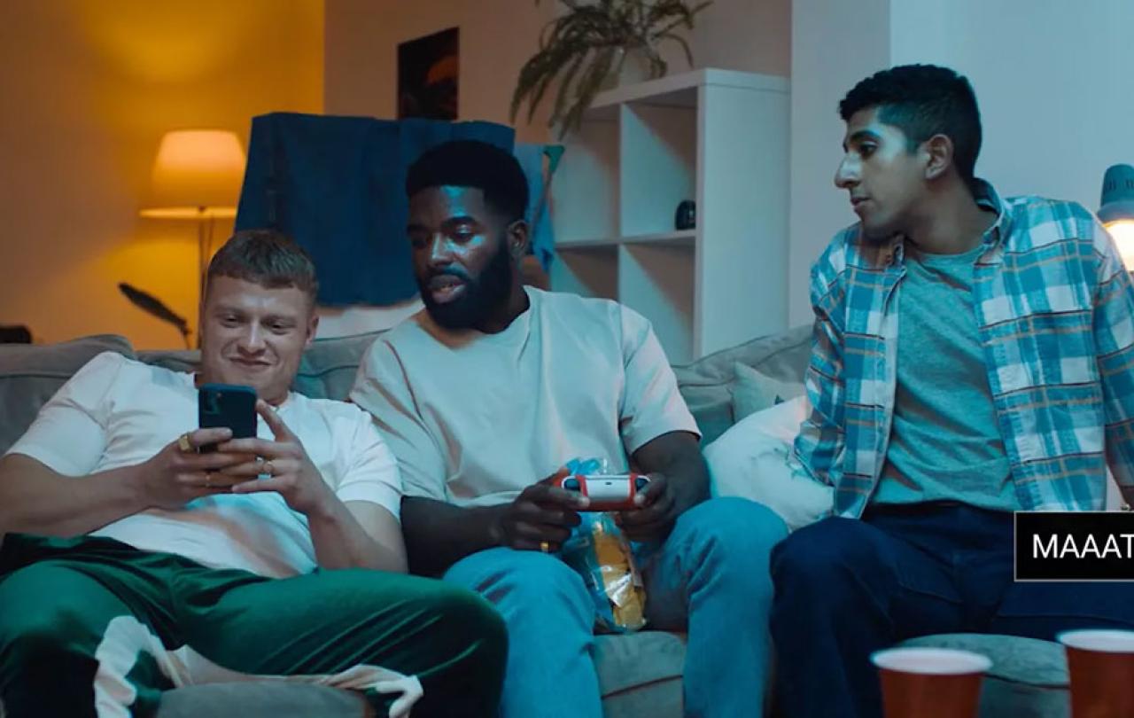 Three young men sit on a couch. One is leering at a phone while the others look on hesitantly