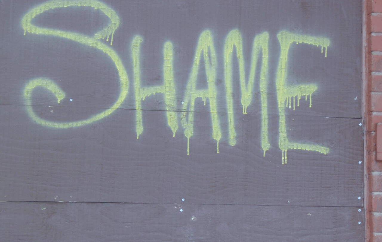 The word 'SHAME' spray painted onto a grey hoarding in lime green paint.