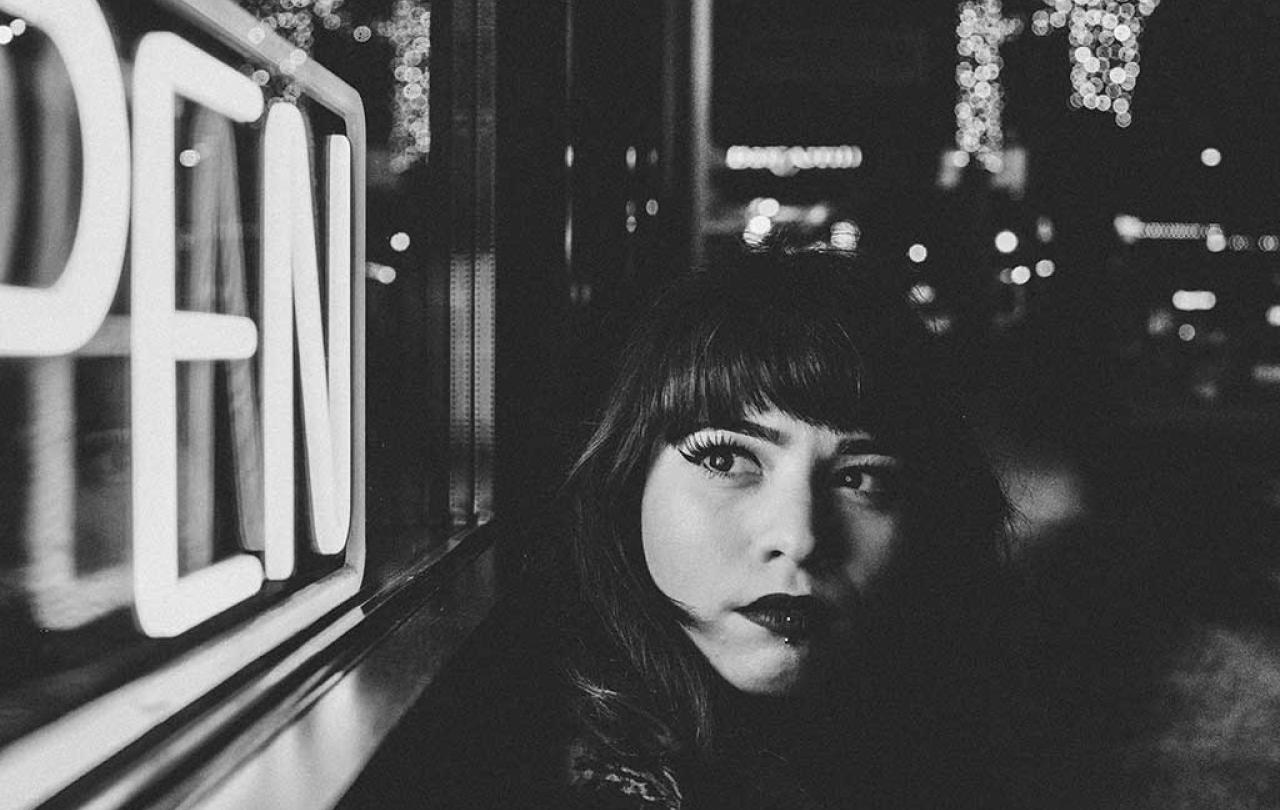 A black and white close up of a women in a street at night, turning to look around at a neion 'open' sign.