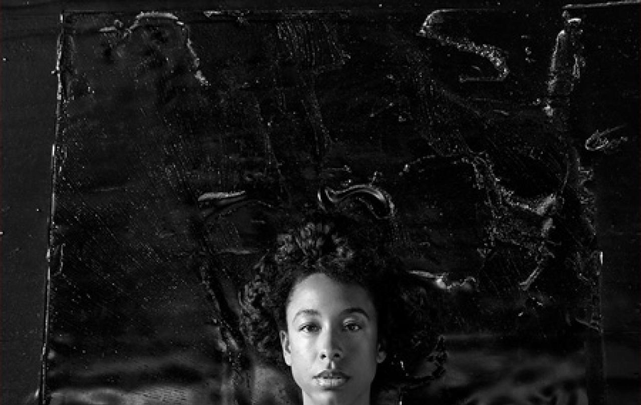 A black and white portrait of a young woman's head against a dark textured background.