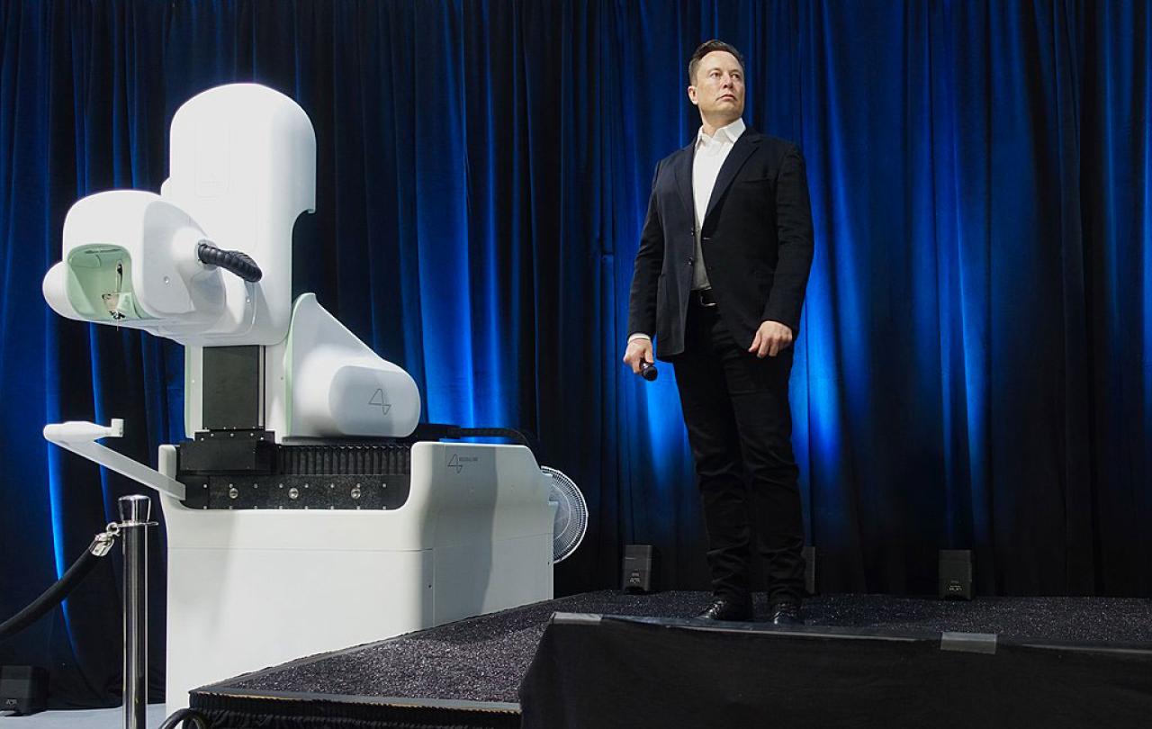 Elon Musk, wearing a dark suit, stands on a stage to a white robotic looking surgical robot.