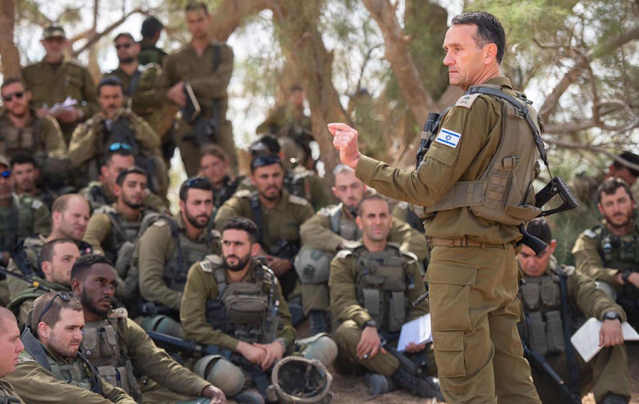 An army general in green fatigues stands and address a group of soldiers sitting, squatting and standing around him