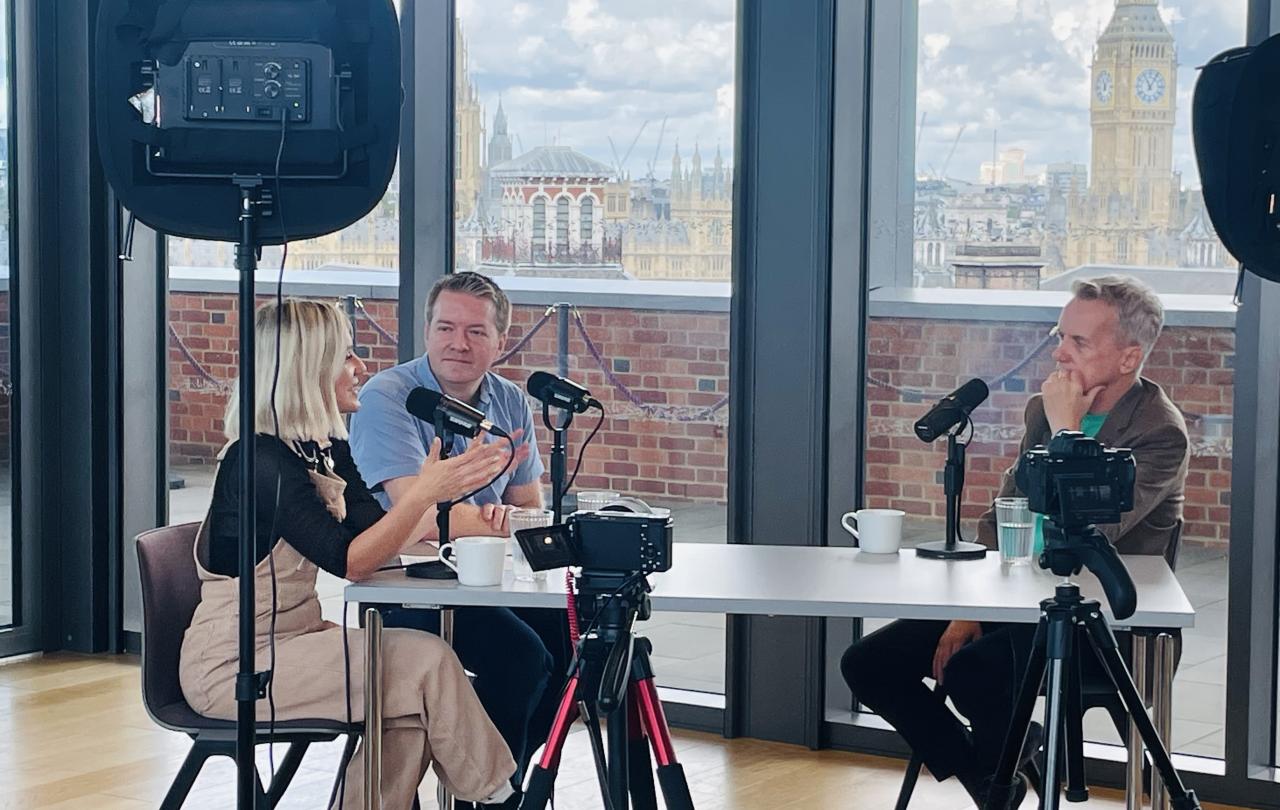 Around a table with microphones, three people record a podcast, one leans in talking and gesturing with a hand while the others listen.