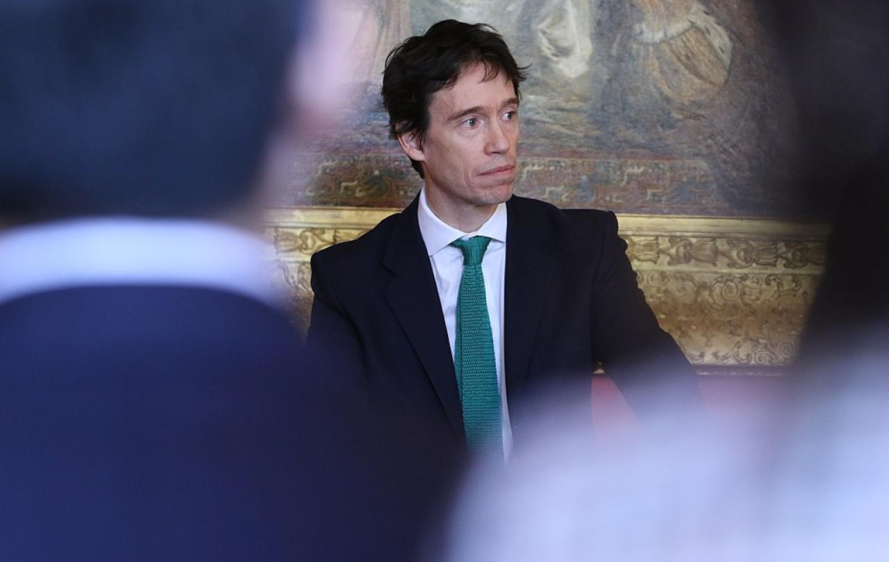 A suited politician stands looking pensive, framed by two out of focus audience members.