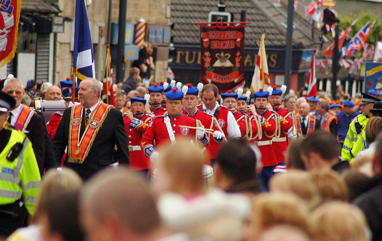 Across the heads of a roadside crowd, men wearing orange sashes and military band uniforms march along.