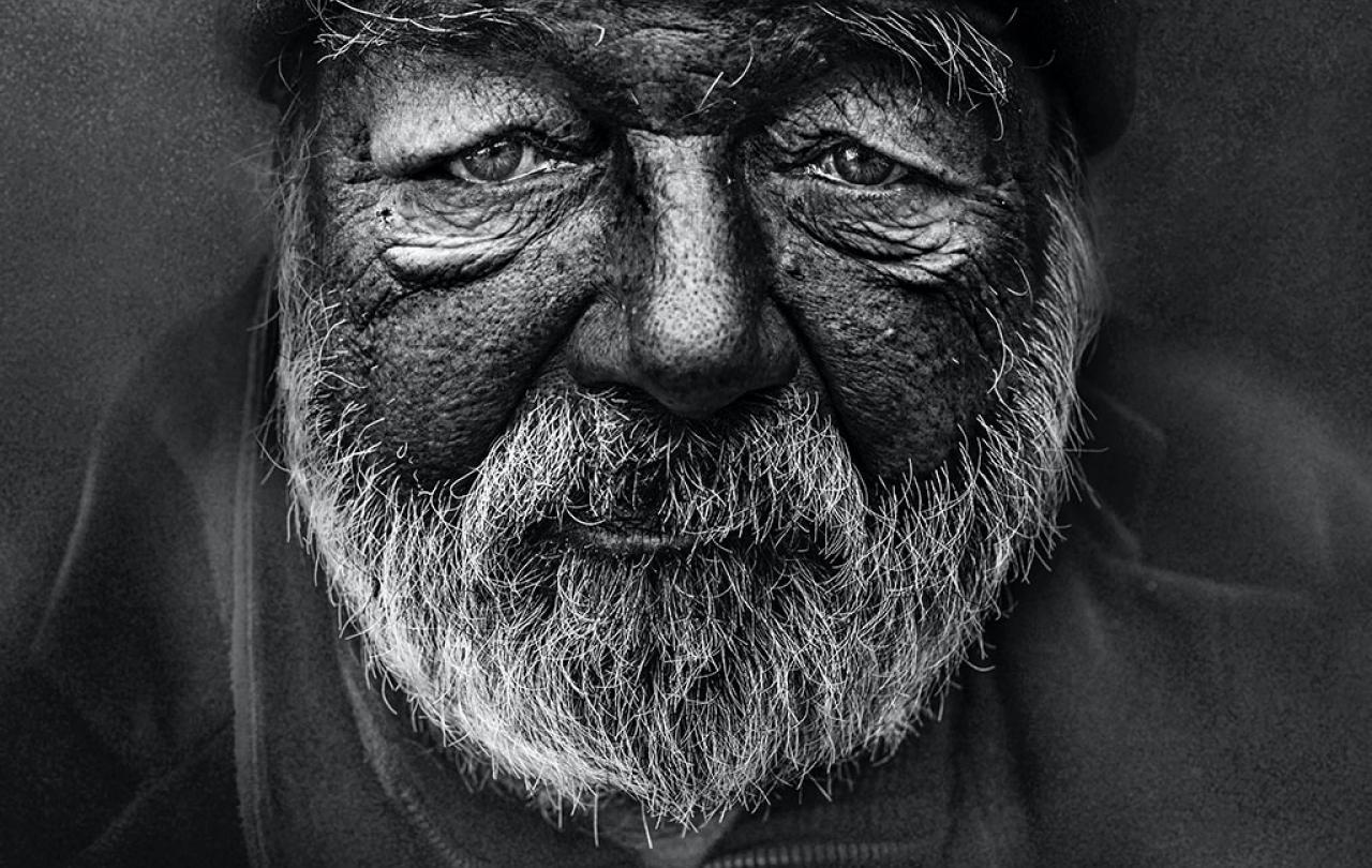 A black and white close up of the weather-beaten and wrinkled face and beard of a homeless man.