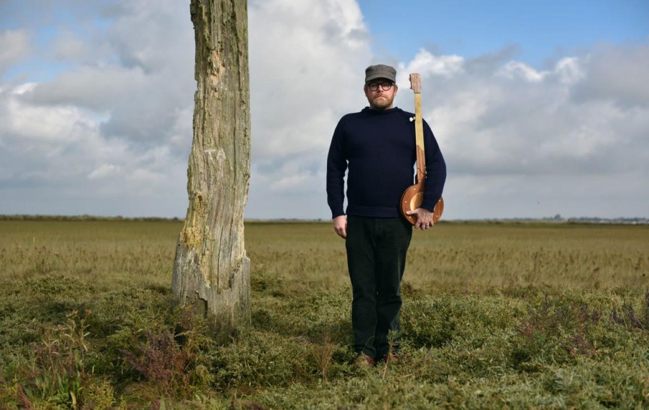 Standing on a salt marsh beside a wooden pillar, a man holds a banjo upright like a rifle.