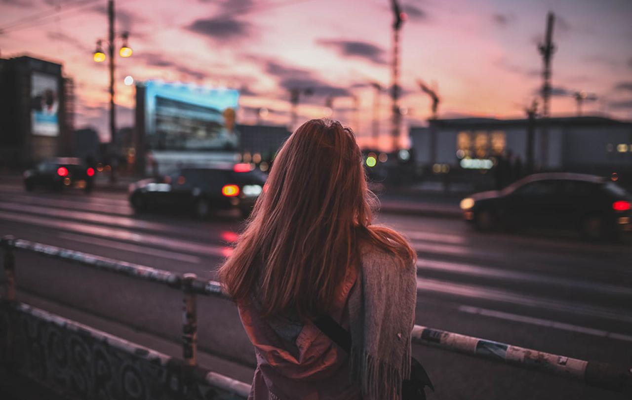A woman stand beside a busy road with her back to the camera. There is a red sky behind her.