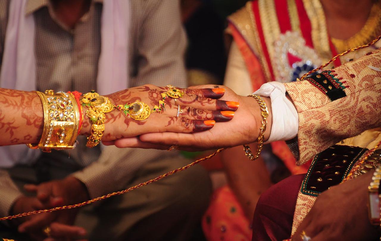A close-up of a bride groom holding the brides hand. Her hand is henna tattooed and bears gold rings and bracelets.