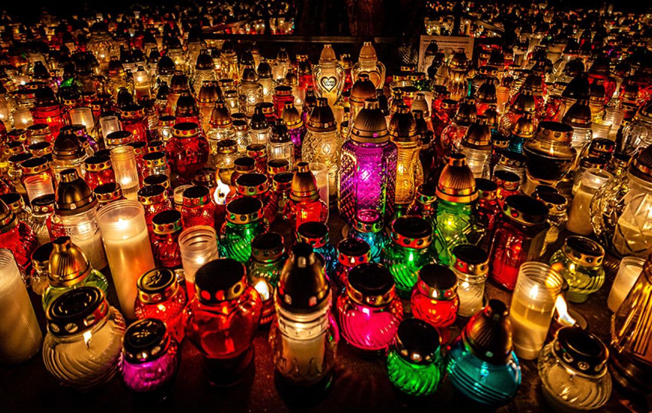 Dozens of candles in cloured jars and holders litter the ground of a cemetry.