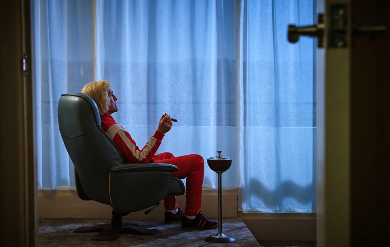 A man in a tracksuit sits in an arm chair smoking a cigar while looking towards a curtained window.