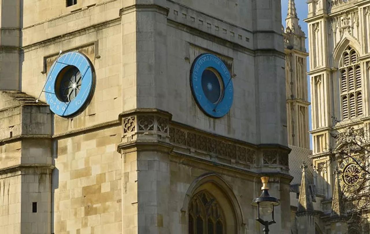 A close up of a blue sun dial on a church clock tower, with an abbey's windows behind it.