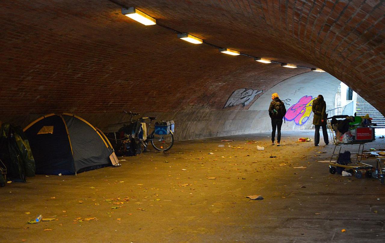 In an underpass a pedestrian passes and look at the tent of a homeless person.