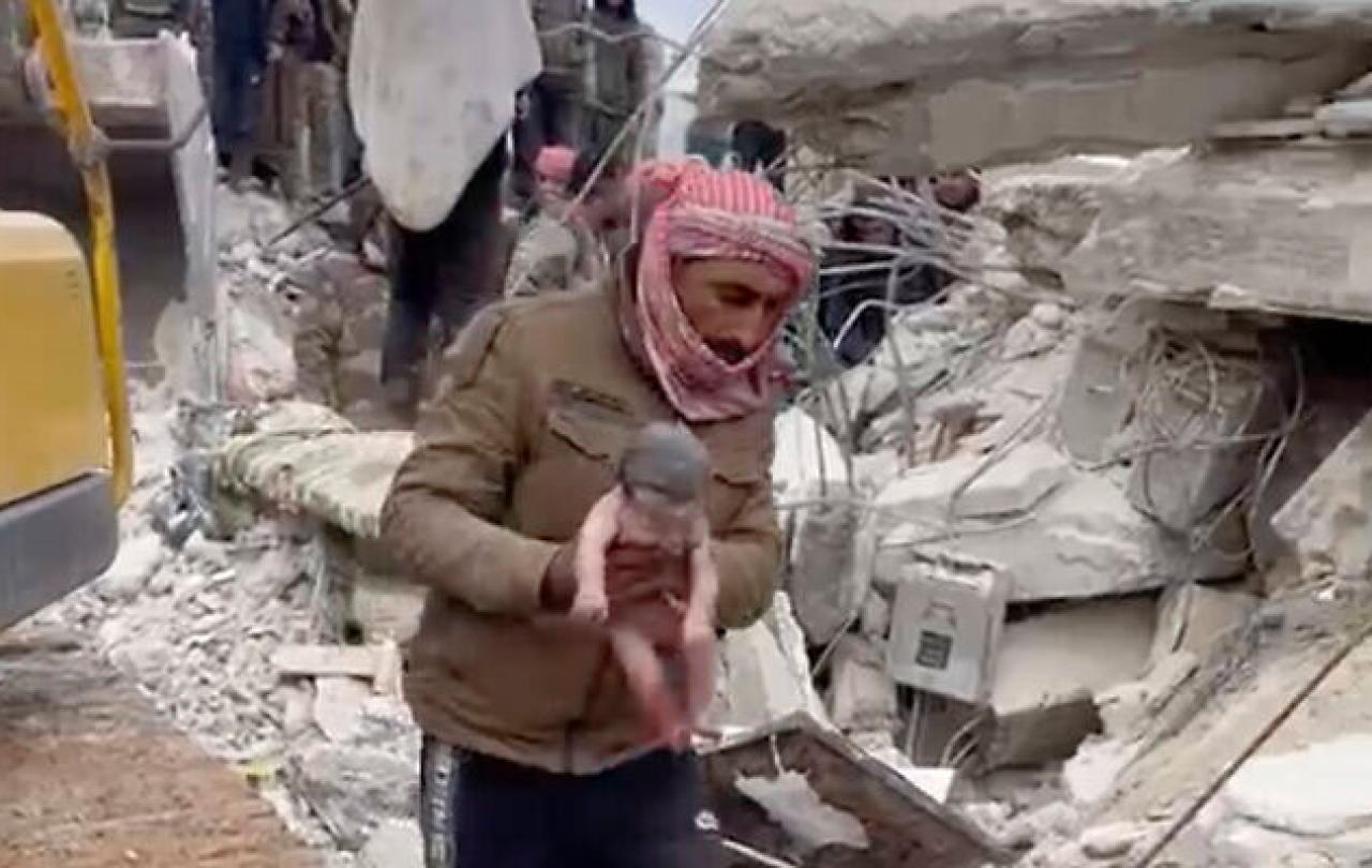 In front of a collapsed building, a rescuer carries a new born baby by the arms.