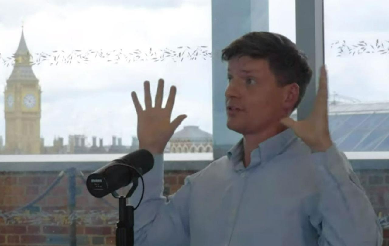 A man sits at a table recording a podcast and holds his hands up for emphasis.