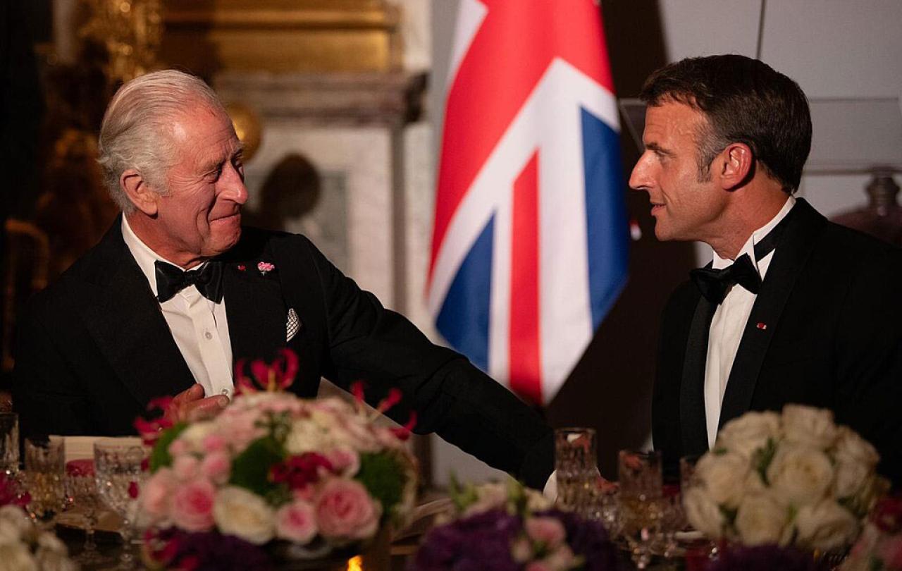 King Charles in a black dinner suit talks and gestures to President Macron who is similarly dressed.