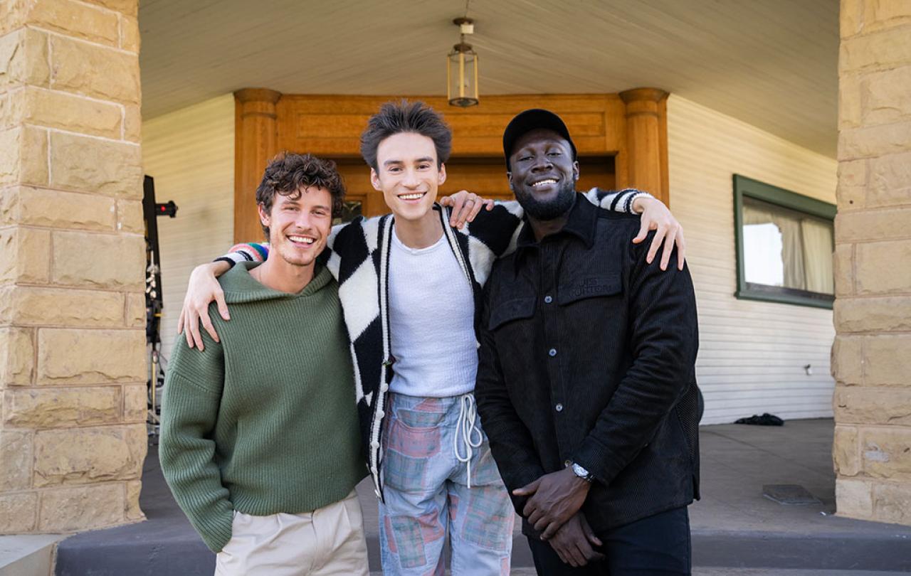 Three happy men stand with the one in the middle draping his arms around his friends.