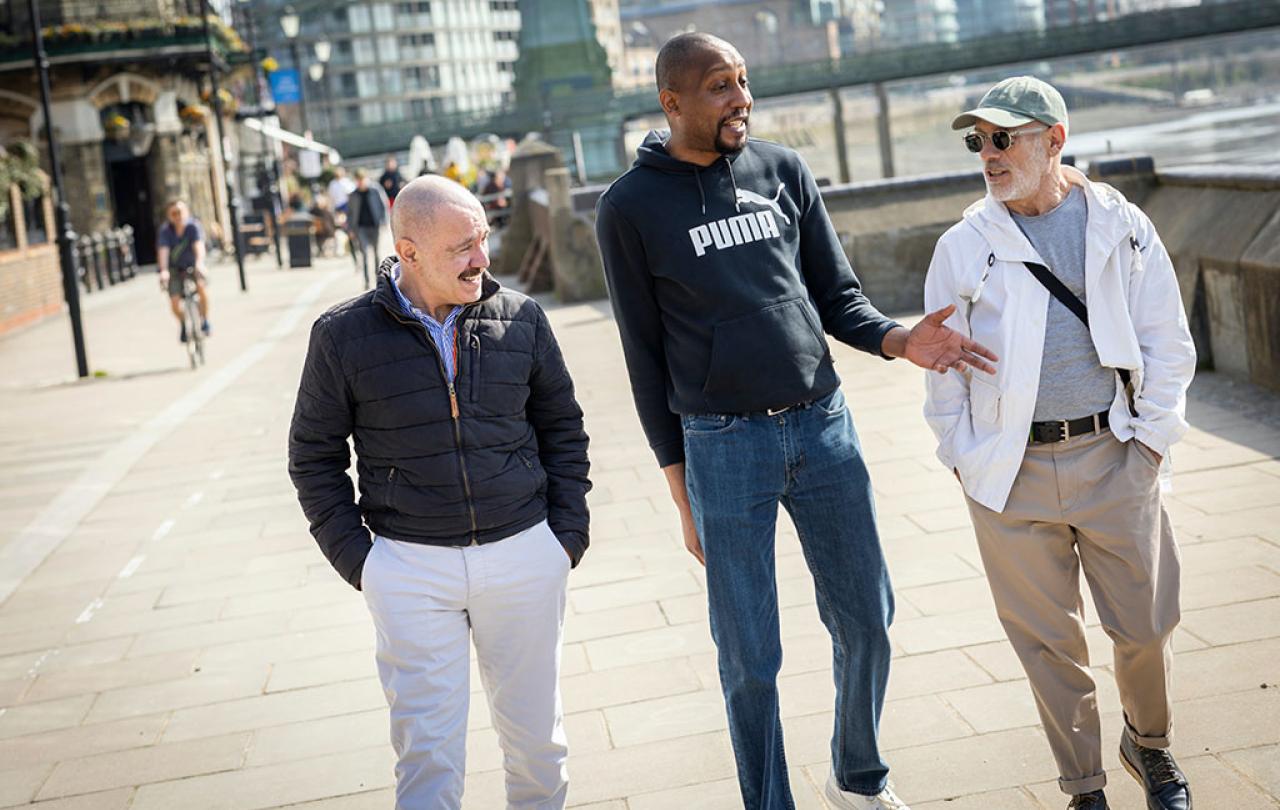 Three men walk down a path, the middle one talking and gesturing while the others listen.